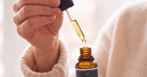 Hand releasing a drop of golden oil from a dropper above an open raw hemp oil bottle by a bright window